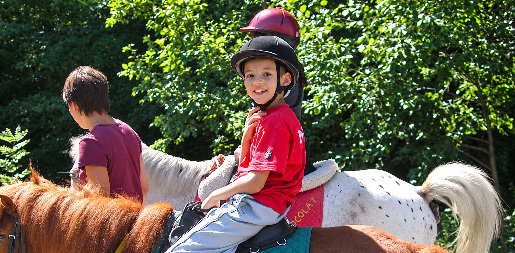 Un jeune garçon en t-shirt rouge et casque noir sourit à la caméra, assis sur un poney blanc tacheté, accompagné d’autres cavaliers dans un décor boisé.