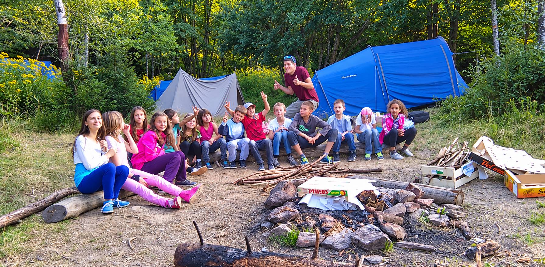 Un groupe d’enfants souriants et un animateur sont assis autour d’un foyer de pierres, devant des tentes bleue et grise en pleine forêt.