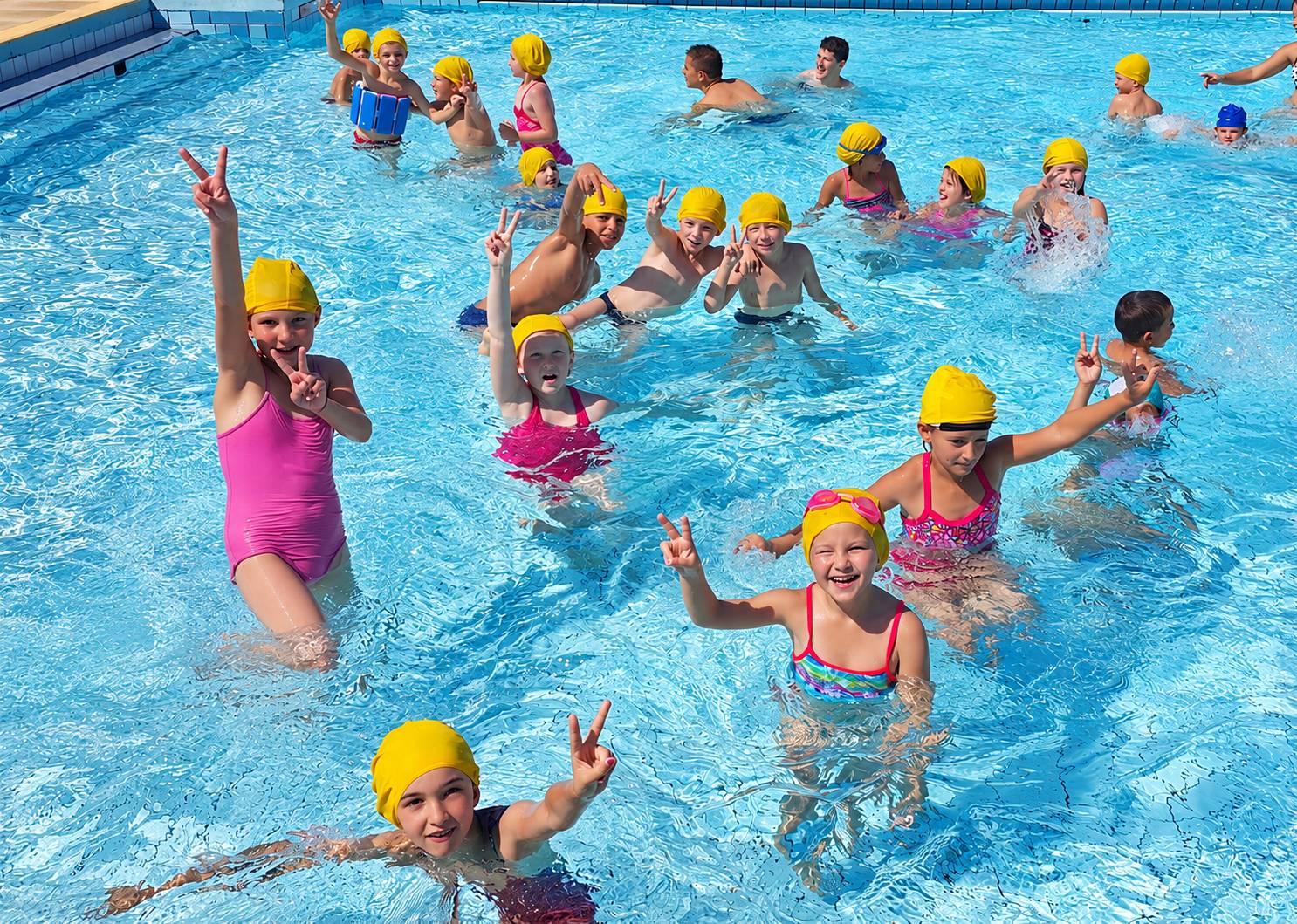 Un groupe d’enfants portant des bonnets de bain jaunes sourit et fait des signes de victoire dans une piscine ensoleillée.