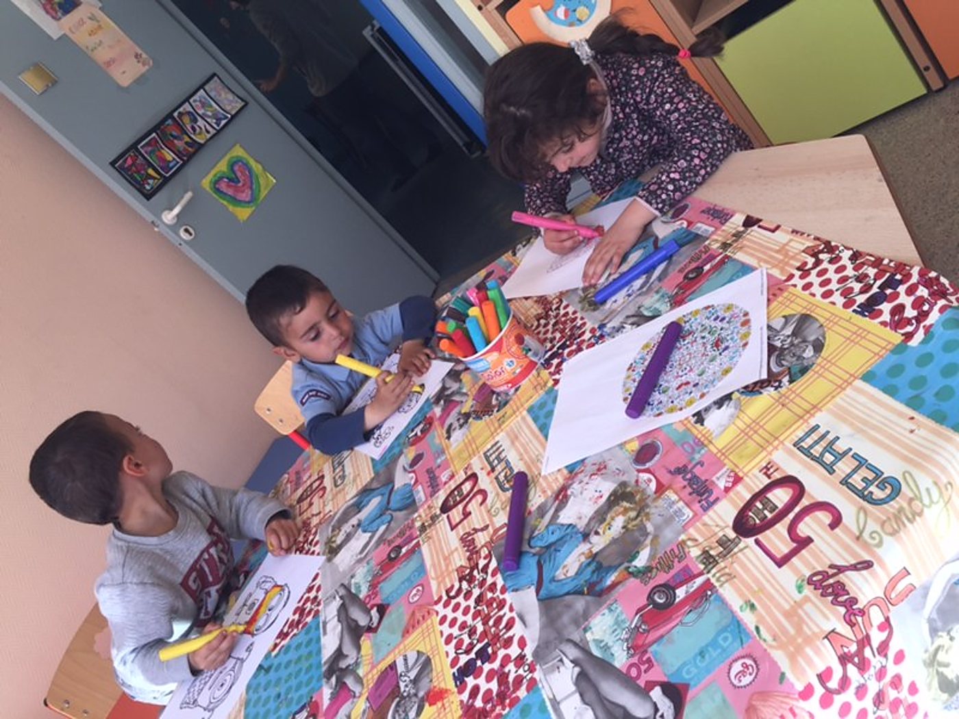 Trois jeunes enfants assis autour d’une table recouverte d’une nappe colorée colorient des dessins avec des feutres dans une salle d’activités.