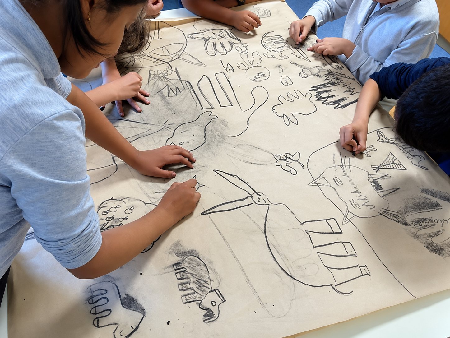 Plusieurs enfants penchés autour d’une grande feuille de papier kraft dessinent des animaux et des motifs au fusain.