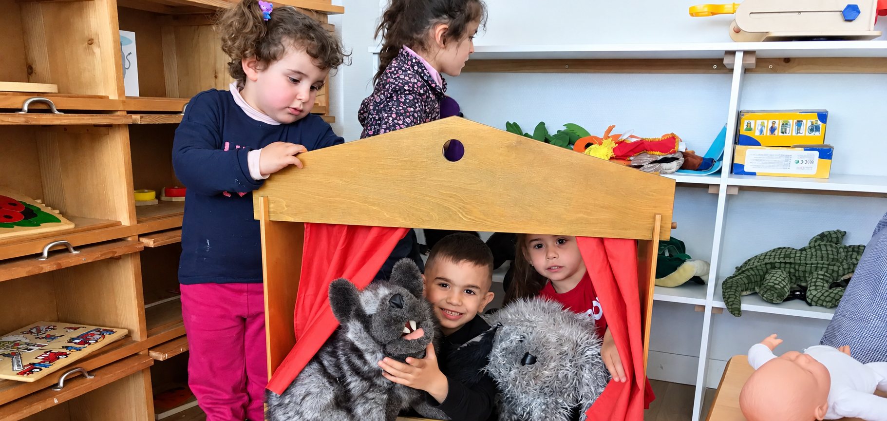 Quatre jeunes enfants jouent autour d’un petit théâtre de marionnettes en bois à rideaux rouges, avec des peluches grises, dans une salle de jeux remplie d’étagères et de jouets.