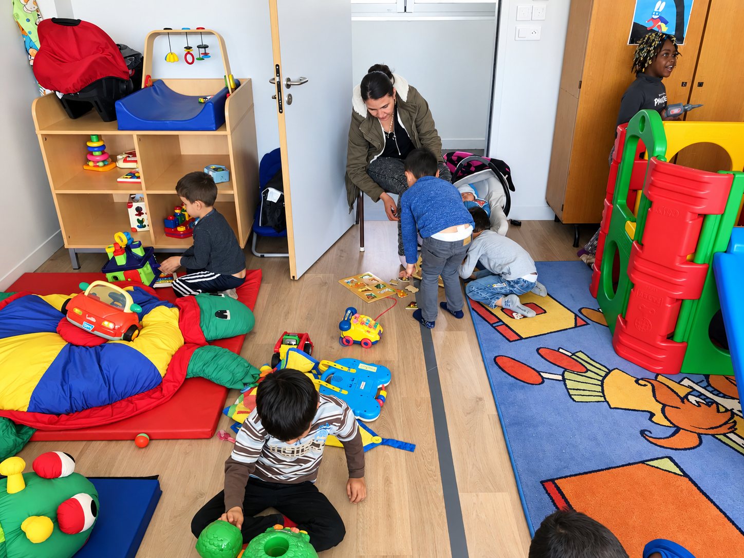 Plusieurs jeunes enfants jouent par terre avec des jouets colorés dans une salle de crèche, sous la surveillance d’une adulte accroupie près d’une porte.
