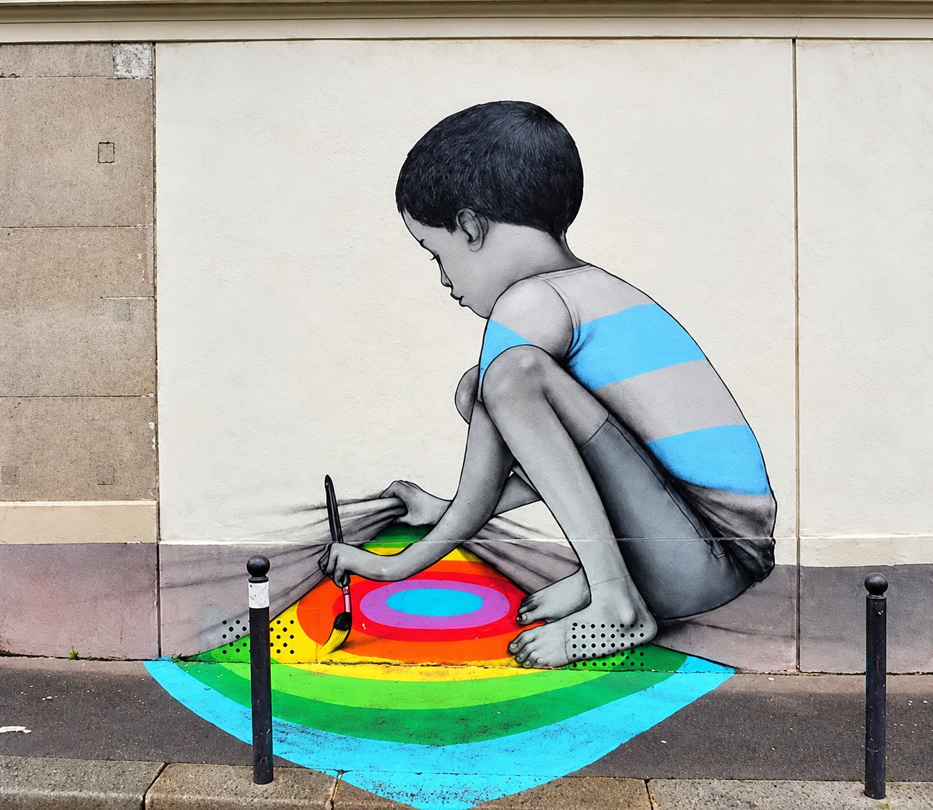 Une fresque en niveaux de gris représente un enfant accroupi qui peint avec un pinceau des cercles colorés d’arc-en-ciel s’étendant sur le trottoir devant un mur blanc.
