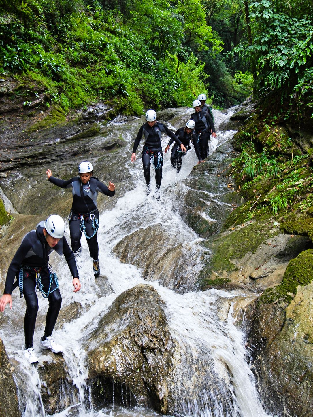 Un groupe de personnes en combinaison néoprène et casque blanc descend à pied un toboggan rocheux d’un canyon, dans l’eau vive entourée de verdure.
