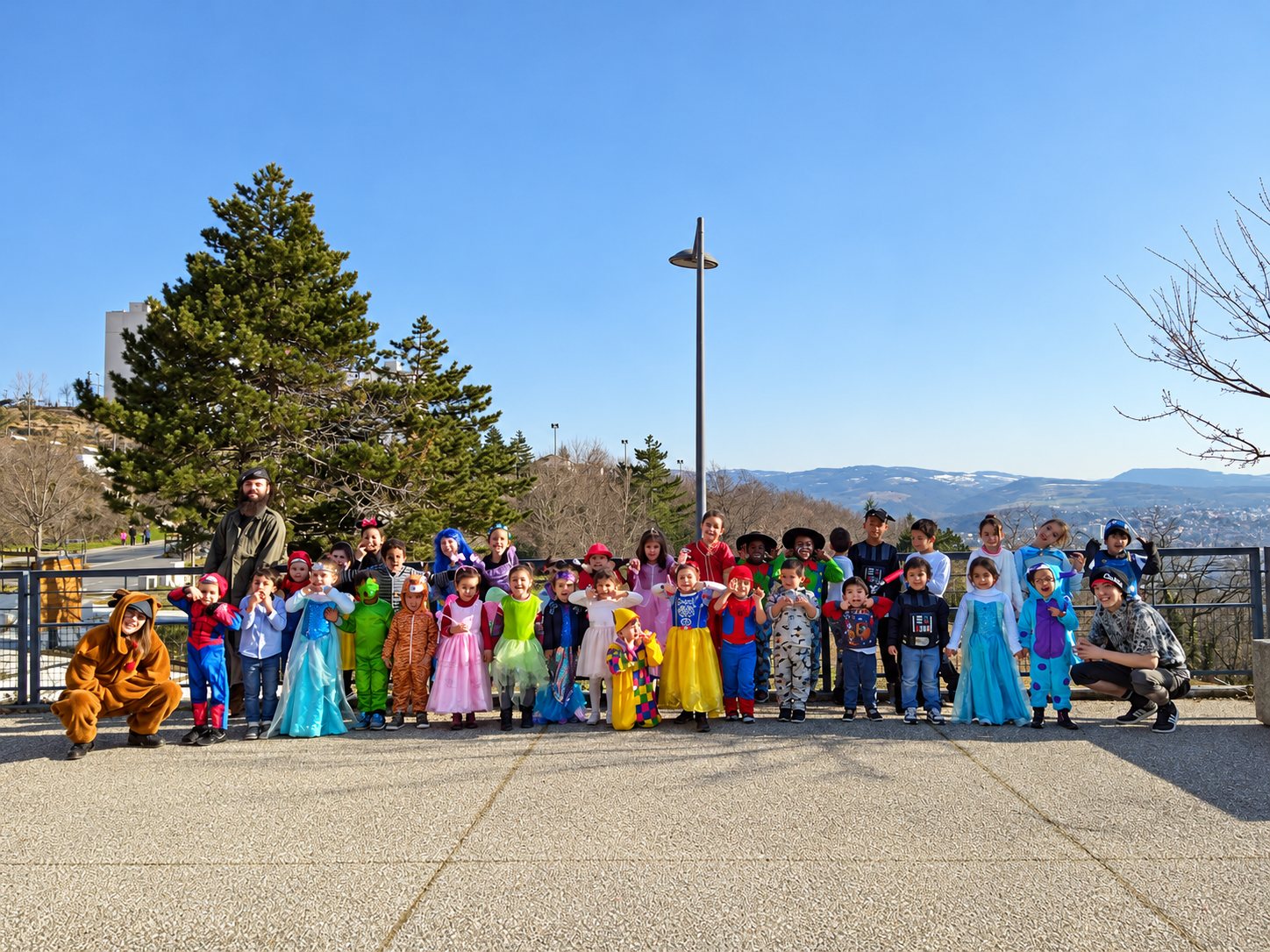 Un groupe d’enfants en costumes colorés pose avec des adultes sur une terrasse en plein air, devant des sapins et des collines.