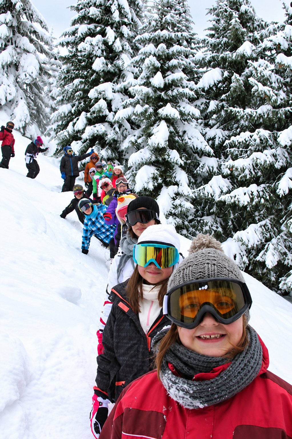 Un groupe d’enfants équipés de lunettes de ski et de bonnets avance en file sur une pente enneigée au milieu de sapins couverts de neige.