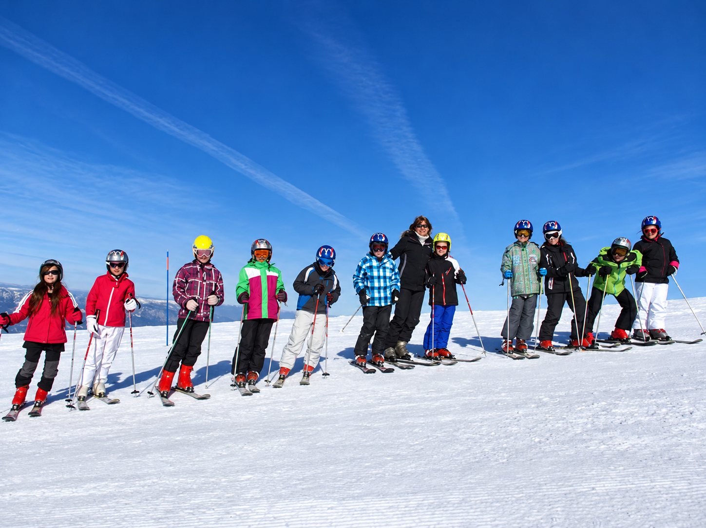 Un groupe d’enfants en tenue de ski et casques pose en ligne avec une monitrice sur une piste enneigée, sous un ciel bleu éclatant.