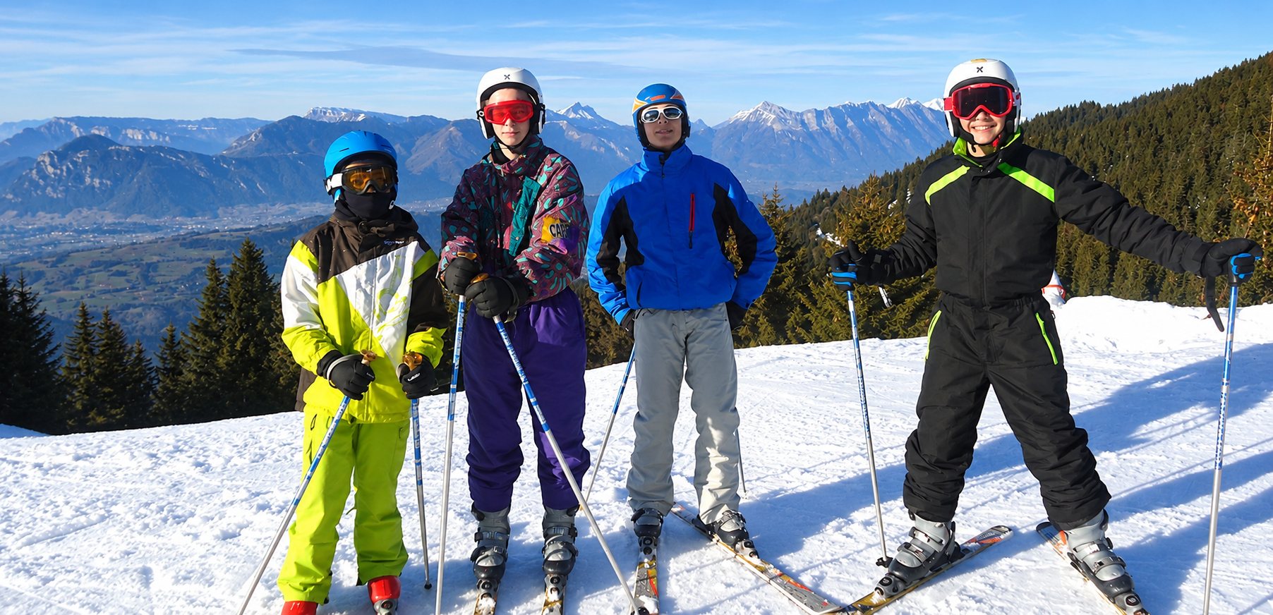 Quatre skieurs casqués et en combinaisons colorées posent sur une piste enneigée avec des montagnes et des sapins en arrière-plan.