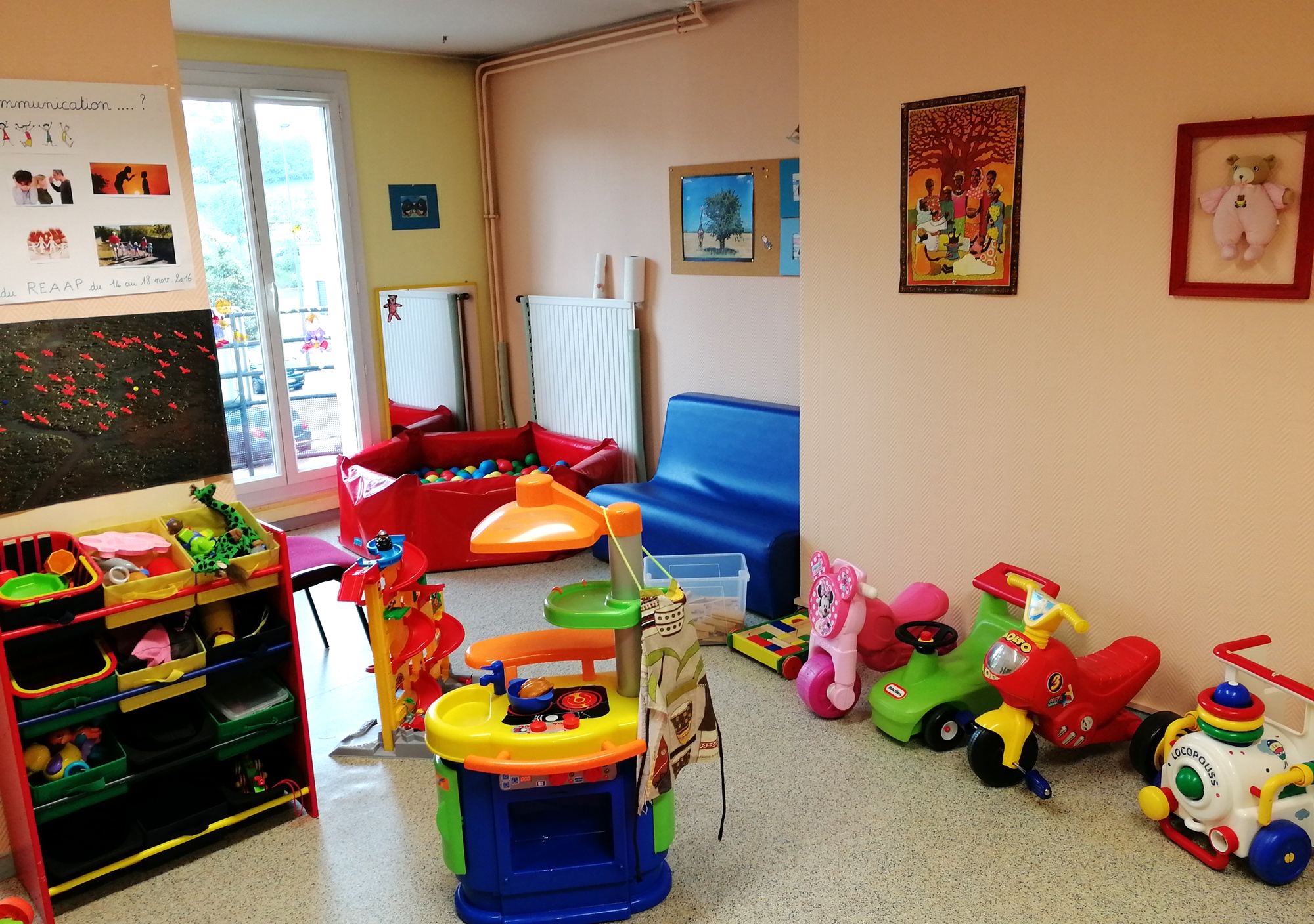 Une salle de jeux lumineuse pour enfants avec une cuisine en plastique, des porteurs colorés, une piscine à balles rouge et une étagère de rangement.