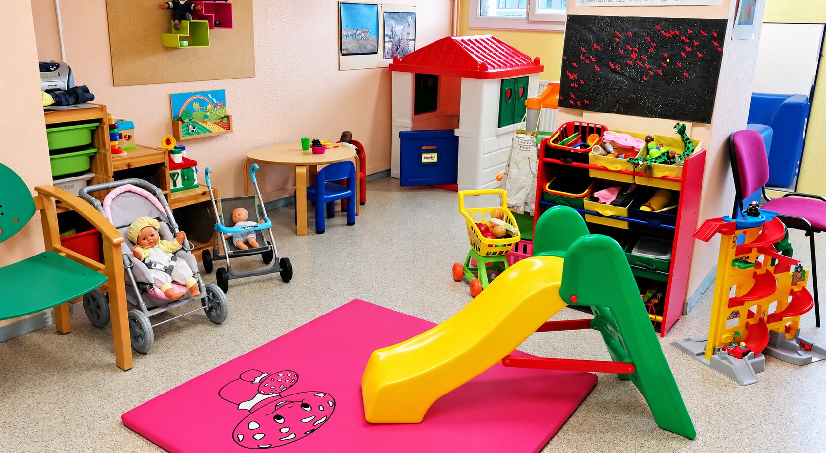 Une salle de jeux colorée pour jeunes enfants avec un toboggan jaune et vert, un tapis rose, des poupées en poussette, une maisonnette et des rangements remplis de jouets.